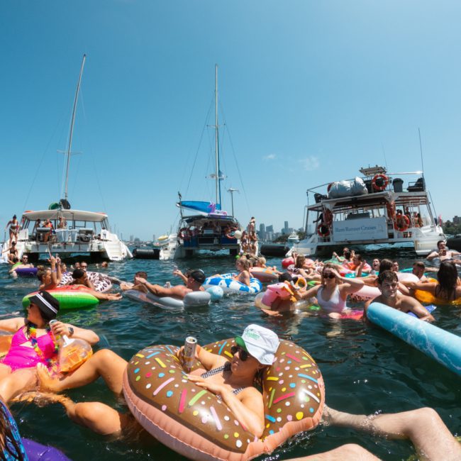 A group of people relax on colorful inflatable floats in the water near multiple boats on a sunny day with a city skyline in the background, enjoying a lively Sydney boat party hire.
