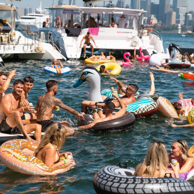 People enjoying a party on the water with inflatable boats and floats, surrounded by larger boats and a cityscape in the background. Various inflatables include a swan, donuts, and others. This vibrant scene is typical of a Sydney boat party hire or private yacht charter Sydney Harbour event.