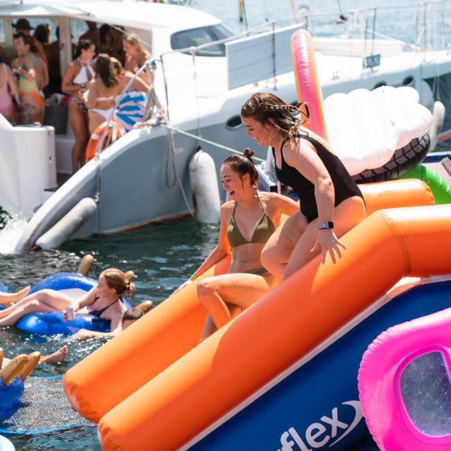 Two women slide into the water from an inflatable slide on a luxurious yacht. Other individuals are seen swimming and relaxing on pool floats nearby, enjoying the perfect day out with DJ boat hire Sydney.