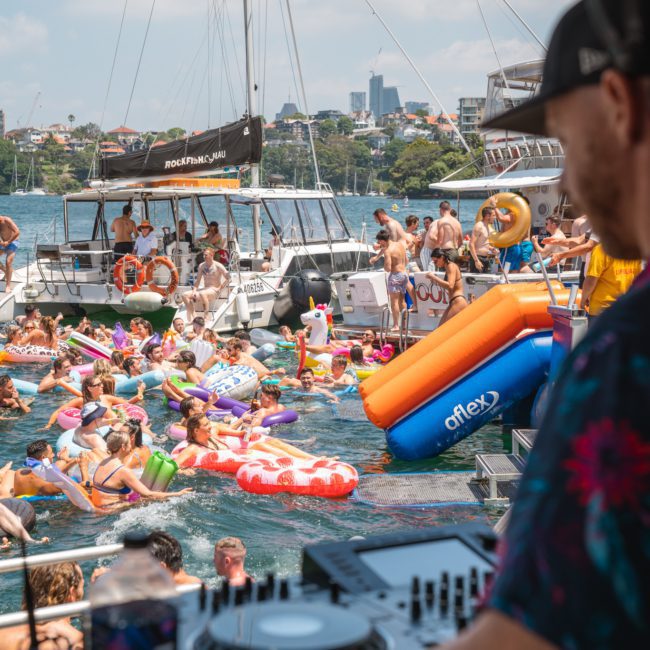 People enjoying a pool party on luxury yachts with inflatables, a DJ playing music, and a city skyline in the background.