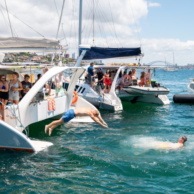 People on anchored boats enjoying a sunny day on the water, with individuals swimming and jumping into the sea. The background shows a distant cityscape and a partially visible bridge, perfect for a thrilling Sydney boat party hire.