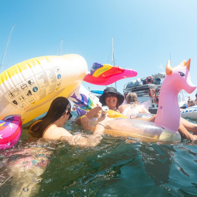 People enjoying a sunny day in the water with colorful inflatable pool floats, near boats in the background, perfect for a catamaran party Sydney.