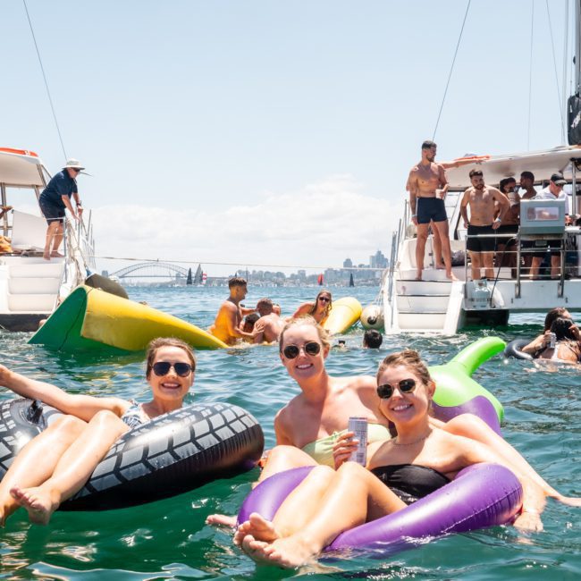 Three people are lounging on inflatable floats in the water, holding drinks. Other people enjoy a Sydney boat party hire on two boats and swim nearby, with a beautiful cityscape visible in the background.