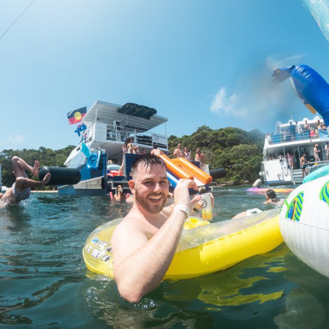 A man smiles at the camera while floating in the water on a yellow inflatable ring. Others are swimming and climbing onto nearby boats in a lively water scene, perfect for a Sydney boat party hire or luxury yacht hire Sydney experience.