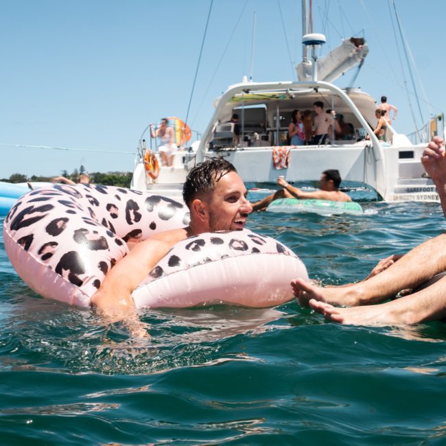 A man floats on a leopard-print inflatable ring in the ocean near a luxury yacht hire in Sydney. Another man’s legs are visible in the foreground, enjoying the serene waters near a boat filled with people.