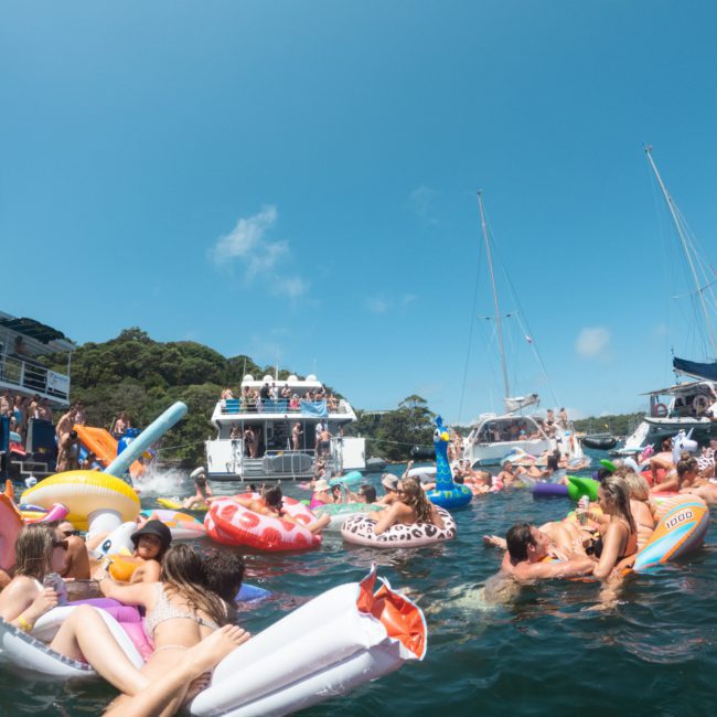 People are floating on inflatables and swimming around boats anchored in a sunny, scenic bay. The water is filled with colorful inflatables, and some people are enjoying a catamaran party in the background.