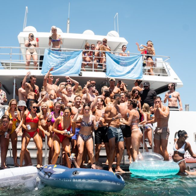 A group of people in swimwear enjoying a private yacht charter on Sydney Harbour with inflatables and drinks, with more people on the yacht’s deck and clear skies in the background.