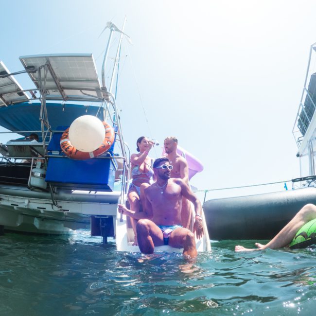 Three people sit on the back steps of a luxury yacht, enjoying the water, while another person floats on a separate inflatable boat nearby. The scene is bright and sunny, perfect for a private yacht charter in Sydney Harbour.