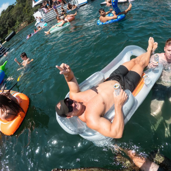 People are enjoying a sunny day in the water, floating on inflatable devices. One person is reclining on an inflatable lounger, holding cans of drinks, while others float and swim around. Imagine adding the elegance of a luxury yacht hire Sydney to enhance this perfect day on the water even further!