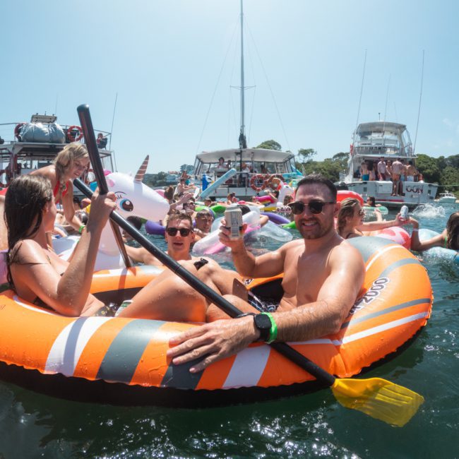People relaxing on inflatable floats in the water, with boats and more individuals in the background enjoying a sunny day. In the distance, a luxurious private yacht charter Sydney Harbour provides an exclusive experience for those aboard.