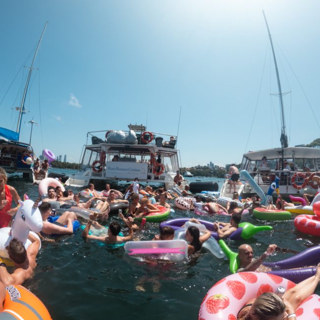 A group of people floating on various inflatable toys in the water near boats on a sunny day, enjoying a Sydney boat party hire.