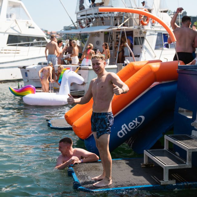 Person standing next to an inflatable slide on a dock, giving a thumbs-up. People in swimwear and floaties are in the water and on boats in the background, enjoying a private yacht charter Sydney Harbour.