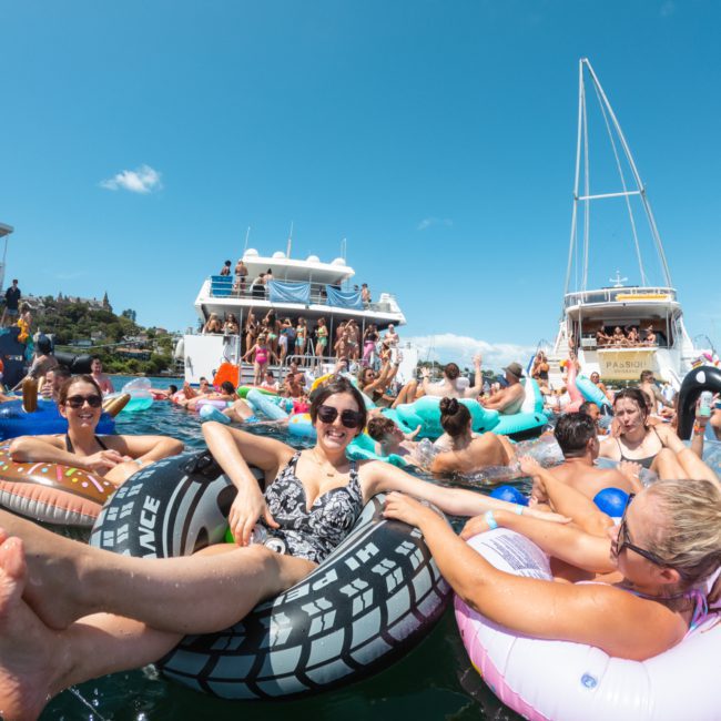 People float in inflatable tubes and lounge in the water near boats under a clear blue sky, enjoying a sunny day at a catamaran party Sydney has to offer.