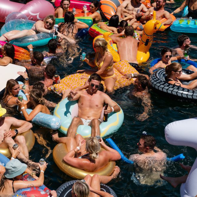 A lively scene of people floating and swimming on a lake, enjoying various inflatable pool toys like unicorn floats and tire rings. Everyone seems to be having a fantastic time in the sunny weather—similar to the fun vibe you'd expect from a catamaran party in Sydney.
