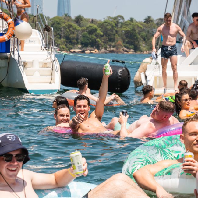 A group enjoys a sunny day on the water, floating on inflatable devices and holding drinks. A luxury yacht hire Sydney is docked nearby, with some individuals aboard. Trees and a building are in the background.
