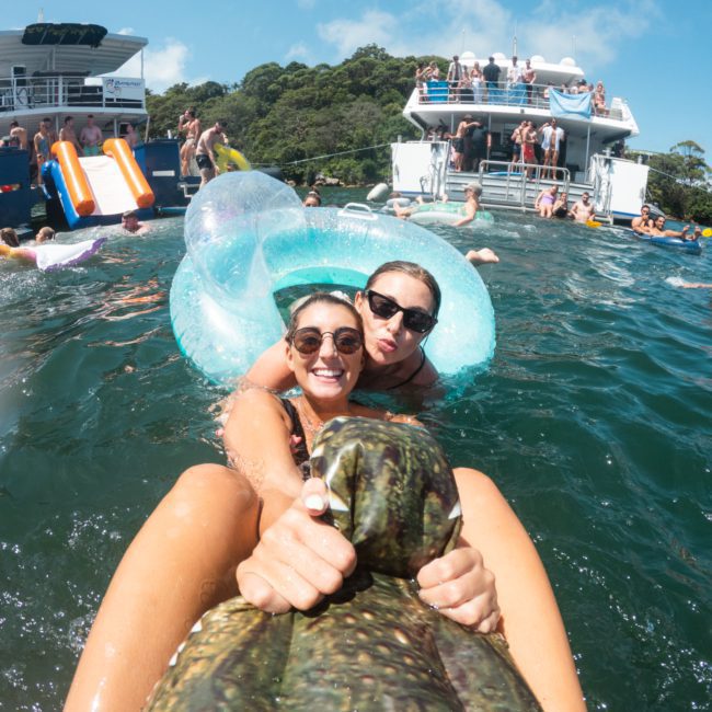 Two women are floating on an inflatable tube in the water near a luxury yacht hire Sydney, surrounded by other people swimming and relaxing.