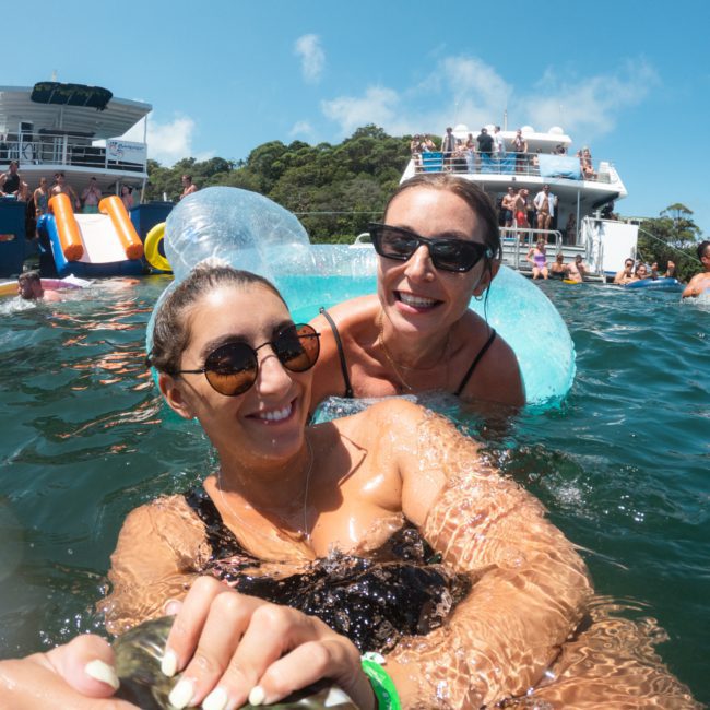 Two women in swimsuits and sunglasses, one in an inflatable ring, are smiling and posing for a selfie in the water. A large boat from a Sydney boat party hire and other people are visible in the background.