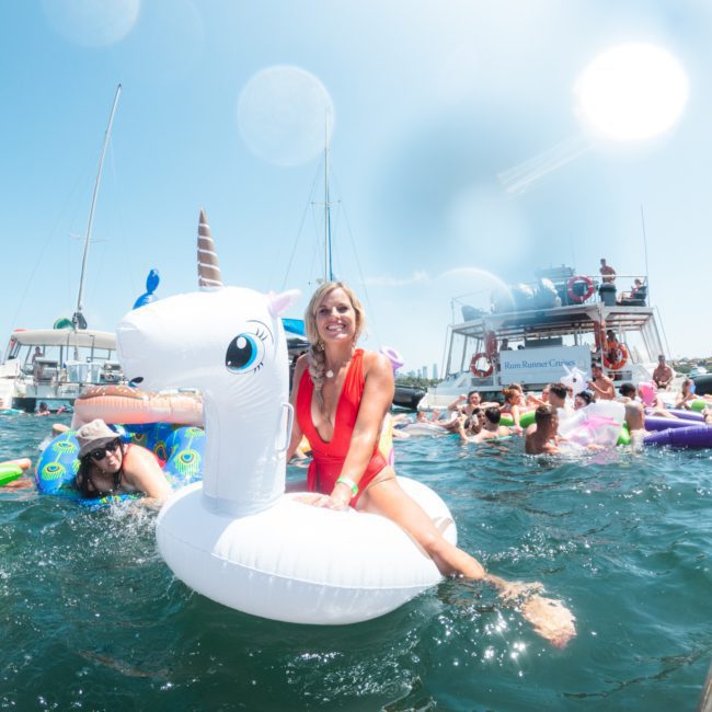 A person in a red swimsuit is floating on an inflatable unicorn in a body of water with boats, including a lively catamaran party Sydney, and other people in the background.