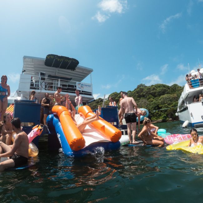 People are swimming and using inflatable floats near boats docked by a forested shoreline on a sunny day, while others enjoy the luxury of a private yacht charter Sydney Harbour.