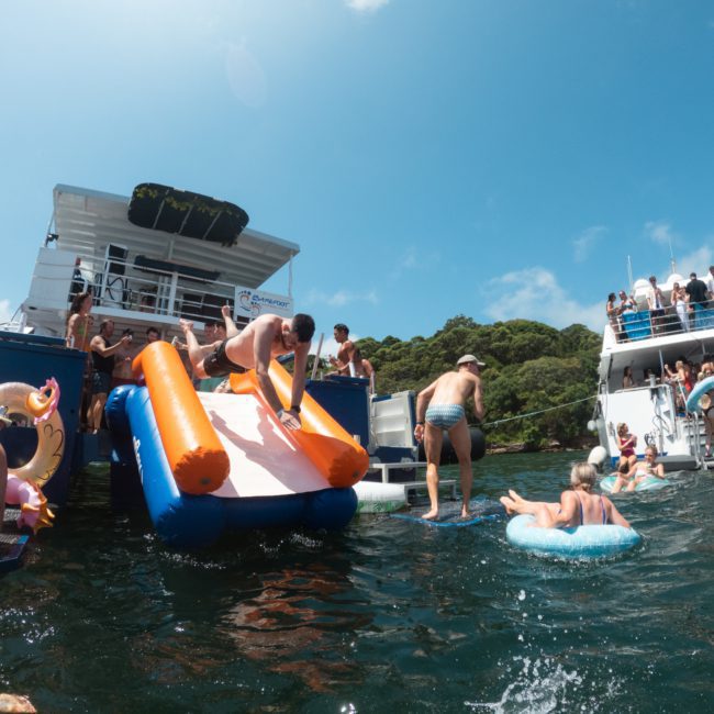 People are enjoying a sunny day on the water with inflatable floats and slides, gathering around two large white boats anchored near a lush, green shoreline. Add to the fun with a DJ boat hire in Sydney for an unforgettable aquatic experience.