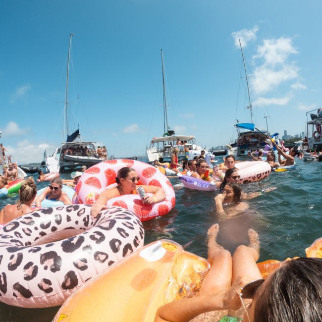 People on inflatable floats in the water surrounded by boats under a sunny sky, enjoying a Sydney boat party hire experience.