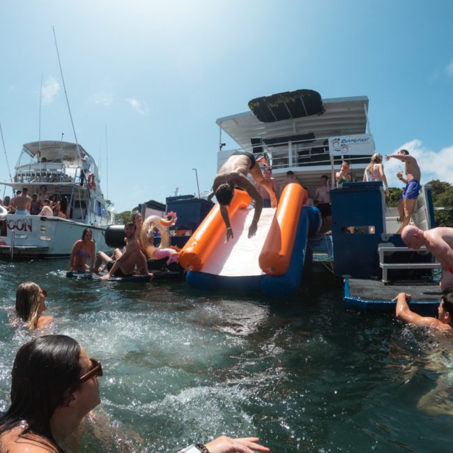 A group of people enjoy the water near boats, with one individual sliding off a water slide from the back of a boat into the sea. Perfect for Sydney boat party hire or luxury yacht hire Sydney, it promises an unforgettable time on the waves.