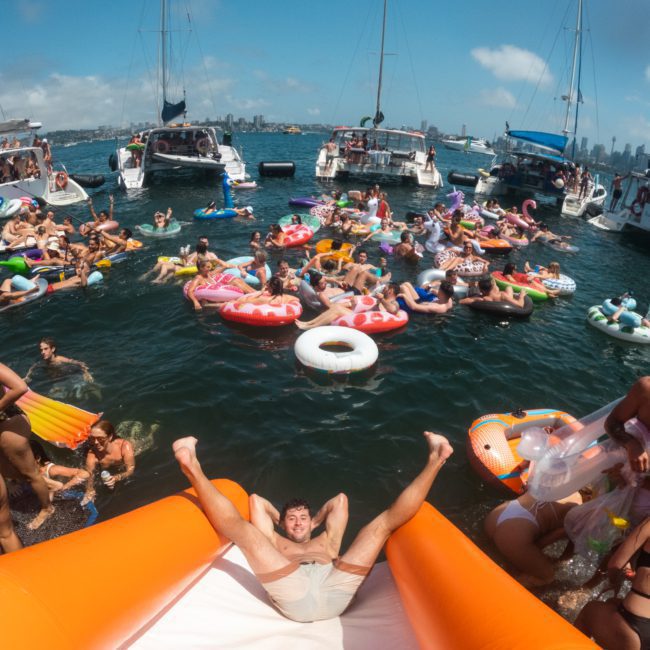 People on various inflatable floats enjoying a sunny day on the water, with multiple boats in the background. A person slides down an orange inflatable slide into the water, while a private yacht charter in Sydney Harbour sails nearby.