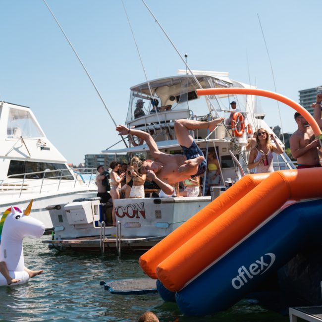 A man performs a backflip off an inflatable slide into the water, while several others enjoy a sunny day on boats during a Sydney boat party hire. A unicorn floaty is seen nearby.