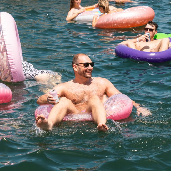 Adults relaxing on colorful inflatable pool floats in a large body of water, enjoying drinks under the sun, could spot a luxury yacht hire Sydney gracefully sailing nearby.