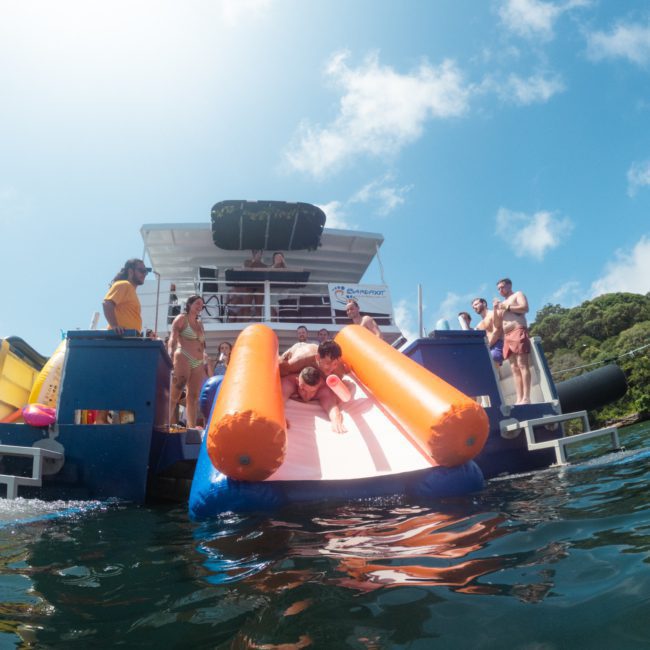 People on a luxury yacht hire in Sydney enjoying the water, with some sliding down an inflatable slide into the sea. Trees and a blue sky are visible in the background.