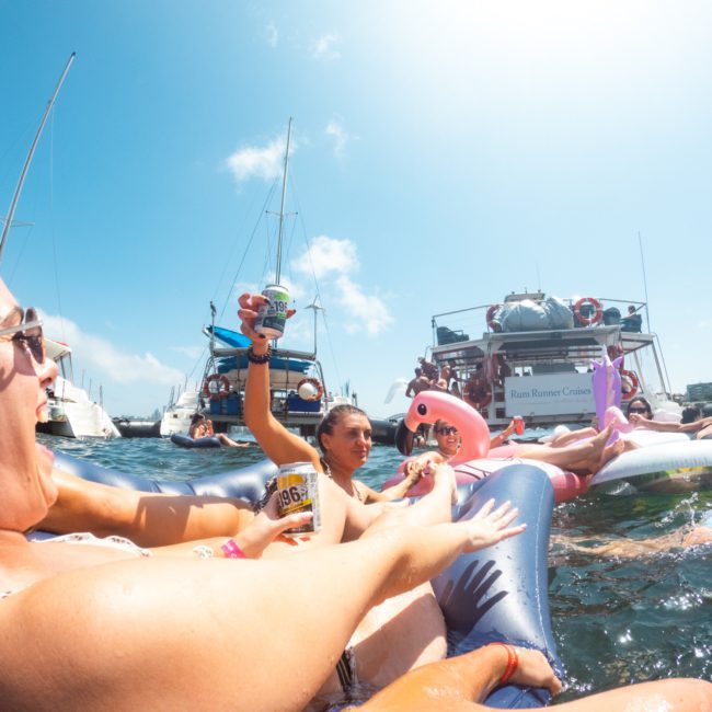 People in swimsuits relax on inflatable floats in the water, holding drinks, with boats in the background on a sunny day. A DJ boat hire Sydney sets the perfect ambiance for this fun and lively scene.