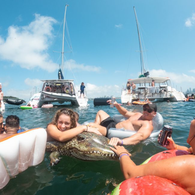 People floating on inflatables in the water with boats in the background on a sunny day. One person is holding a large inflatable crocodile, enjoying a Sydney boat party hire event.