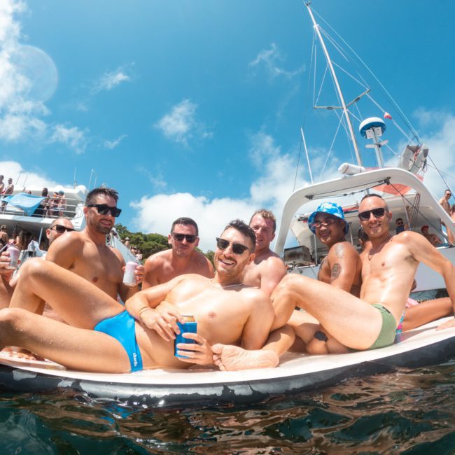 Group of men in swimwear sitting on a boat, smiling at the camera, with a crowd and another boat in the background, enjoying a lively Sydney boat party hire.