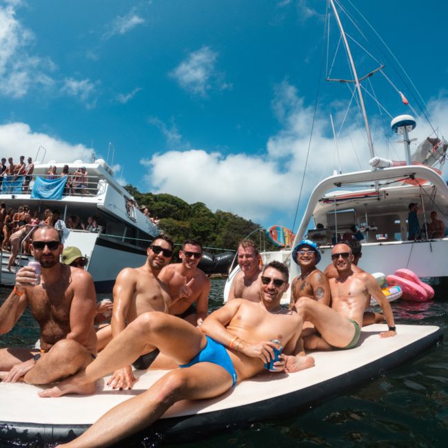 A group of people relax on a floating platform in the water. Boats are anchored nearby, and more people are seen on the boats and in the water. It's a sunny day with a clear blue sky, perfect for enjoying corporate boat events in Sydney.
