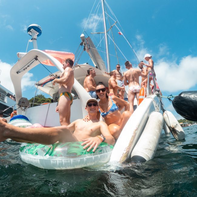 A group of people in swimwear relaxing and socializing on a boat and in the water, with a large sailboat in the background under a clear blue sky, perfect for a Sydney boat party hire or catamaran party Sydney.