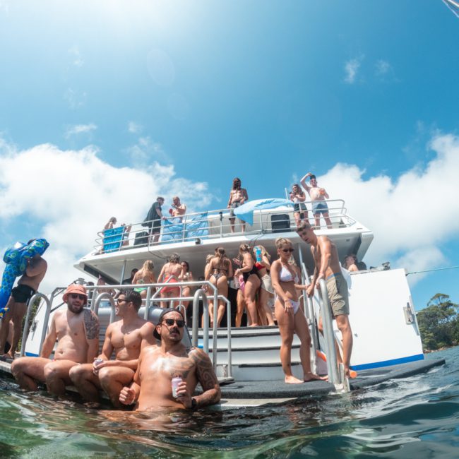 A group of people are socializing and relaxing on a two-level boat and in the water near the stern. The sky is clear, and the scene appears to be a casual gathering or party, perfect for a Sydney boat party hire.