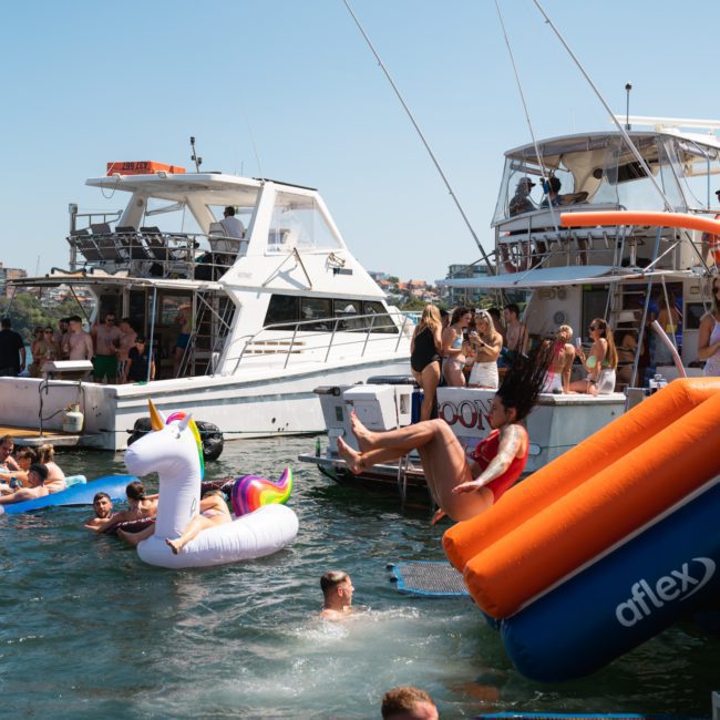 People enjoy various water activities, including using an inflatable unicorn float, near two boats docked close together. Some individuals swim while others relax on the boats during a Catamaran party Sydney event.