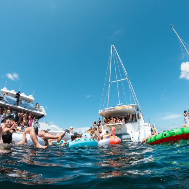 People enjoying a sunny day on the water with various boats and colorful inflatables. Some are swimming, while others aboard a catamaran party in Sydney socialize and relax. The sky is clear with some scattered clouds.
