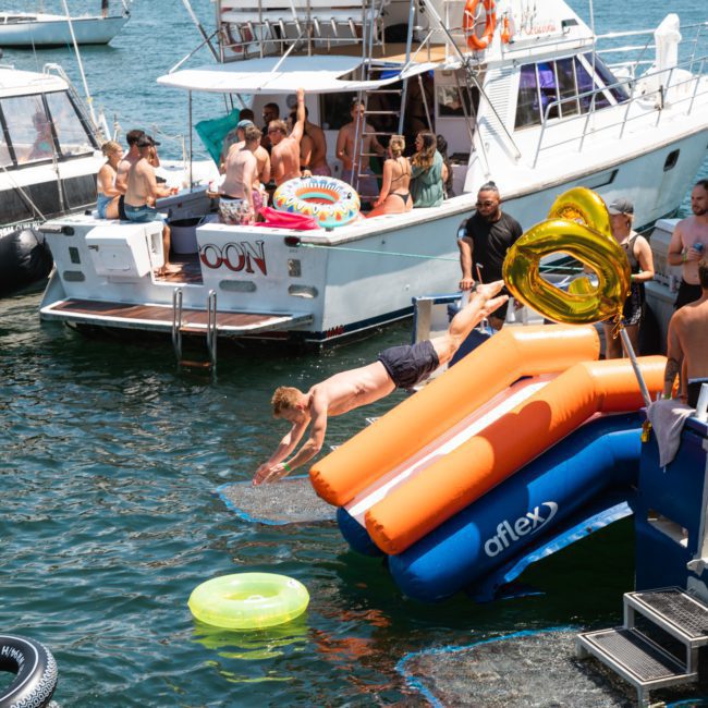 People are gathered on and around a luxury yacht docked in the water, with inflatable floats nearby and someone diving off an inflatable slide into the water.