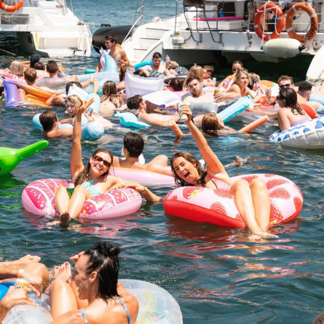 A group of people in an ocean swim and float on inflatable tubes near boats. Two women in the foreground smile and hold up drinks while sitting on inflatable rings, enjoying their time during a luxury yacht hire Sydney experience.