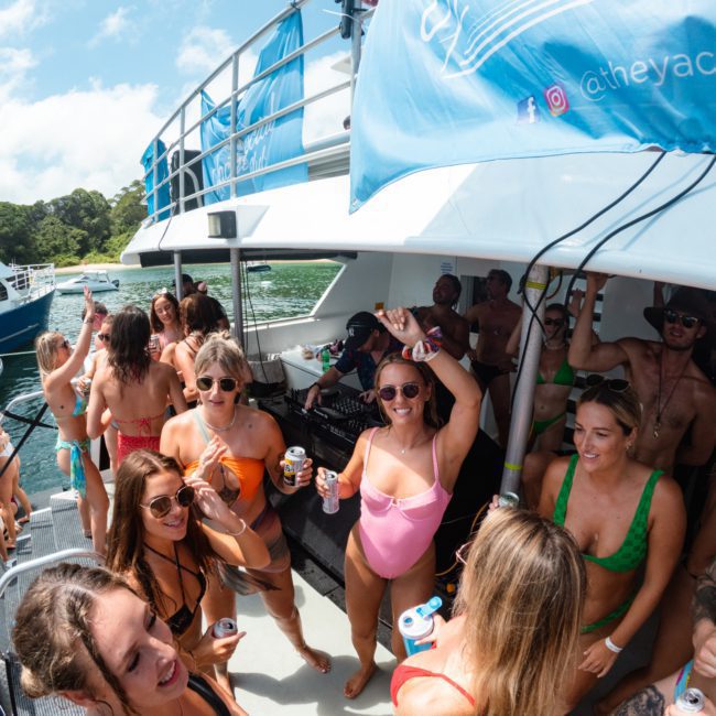 A group of people in swimsuits are having a Sydney boat party hire on a catamaran under sunny weather, holding drinks, and socializing.