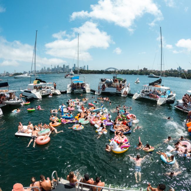 A large group of people relax on colorful pool floats in the water, surrounded by multiple boats on a sunny day with a city skyline in the background, enjoying what looks like an amazing catamaran party Sydney.