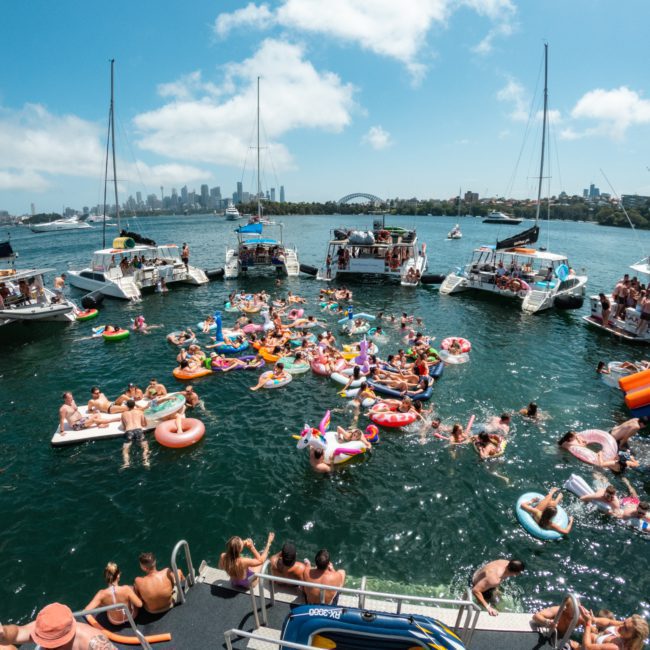 A large group of people on inflatables and boats enjoy a sunny day on the water, with a cityscape and bridge visible in the background. Perfect for a Private yacht charter Sydney Harbour.
