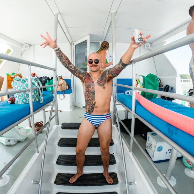 A man with tattoos, wearing striped swimming trunks and sunglasses, stands on boat steps with arms outstretched, holding a drink. A lively crowd and various items are seen in the background, exemplifying the vibrant energy of catamaran party Sydney.