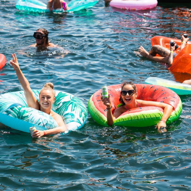 People relaxing on colorful inflatable rings in water, with two individuals in the foreground raising their hands and smiling during a private yacht charter Sydney Harbour.