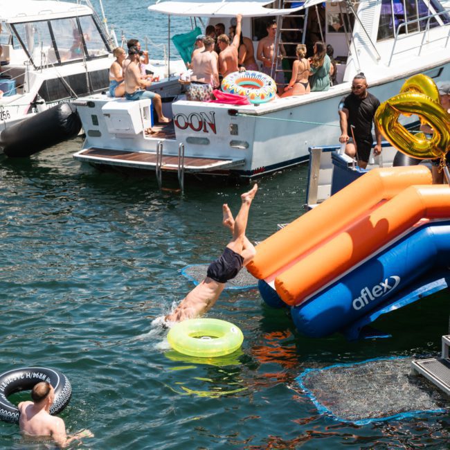 People enjoy a sunny day on the water, with several boats docked. One person dives from a boat into the water using an inflatable slide, while others swim or relax in the boats and water during a luxurious yacht hire Sydney experience.