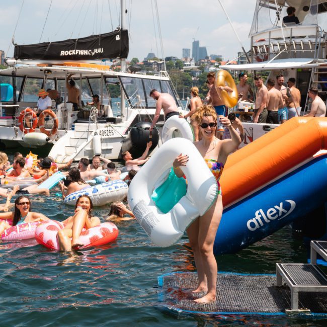 People enjoying a sunny day on a lake with inflatables, floating near boats and a DJ boat hire Sydney. One person stands on a dock holding an inflatable swan.