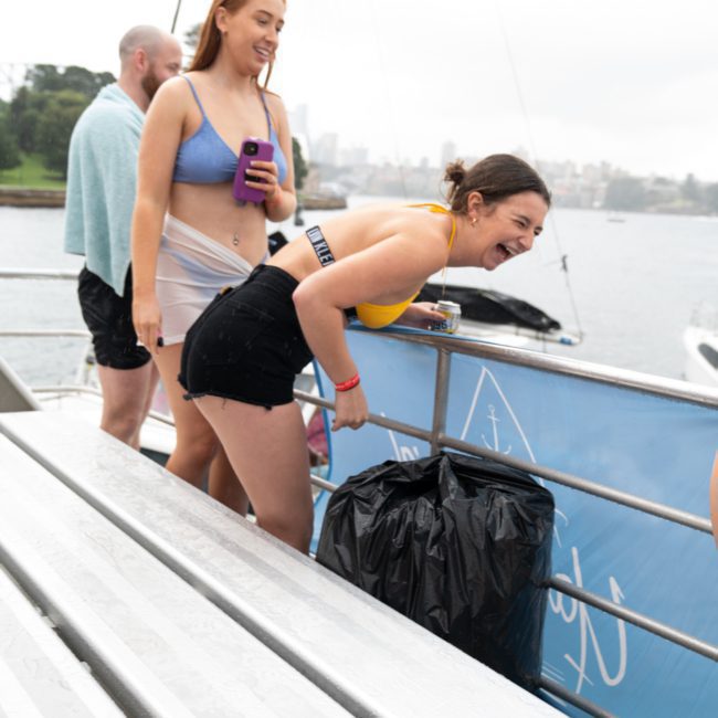 Three people in swimwear standing on a boat dock; two women are smiling and leaning on a railing while a man stands nearby, wrapped in a towel, enjoying the vibe of a DJ boat hire Sydney.