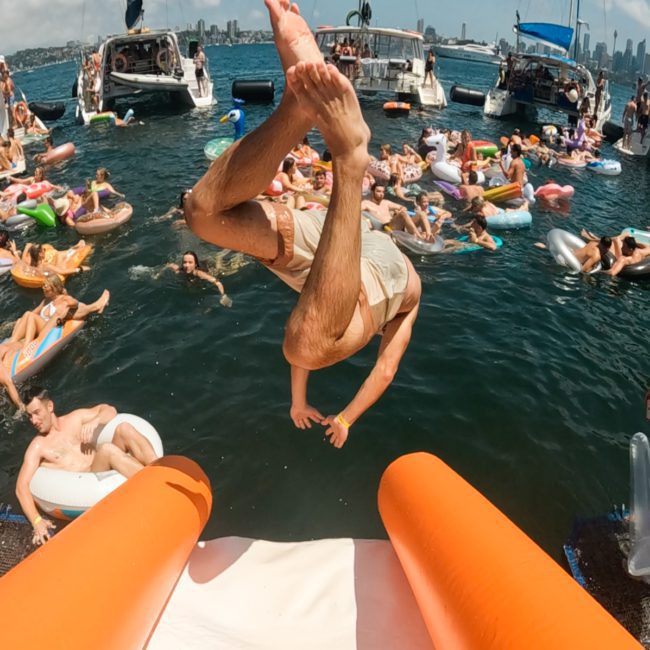 A person mid-flip off an inflatable slide into a crowded body of water with boats and people on inflatables in the background, perfect for a lively catamaran party in Sydney.