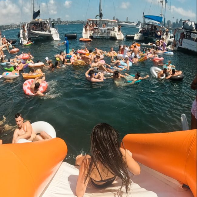 A person is about to go down an inflatable slide into the water, where numerous people are floating on inflatables near moored boats on a sunny day during a lively catamaran party in Sydney.